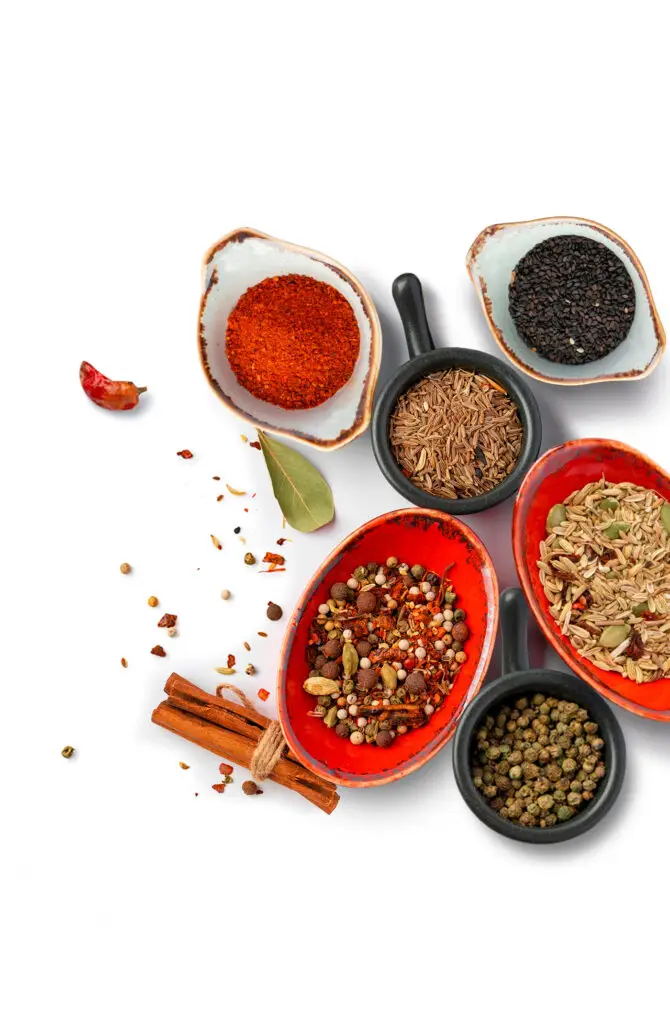 Assorted spices in small bowls on a white background, including black sesame seeds, cumin seeds, fennel seeds, chili powder, and mixed whole spices. A bay leaf, peppercorns, and cinnamon sticks are scattered around.