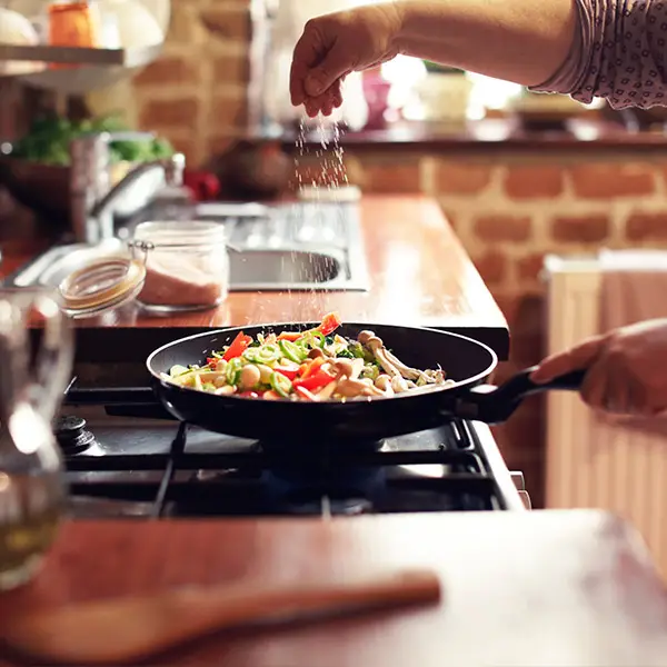 A person sprinkles salt onto a frying pan filled with colorful vegetables on a stove. The kitchen has a rustic look with brick walls and wooden countertops. A jar of salt is open nearby.