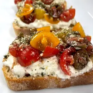 Three slices of toasted bread topped with creamy white cheese, chopped red and yellow tomatoes, herbs, Pesto Seasoning, and a sprinkle of grated cheese are arranged in a row on a white plate.